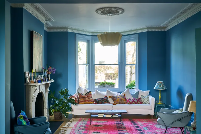 A cozy blue living room with a plush sofa, accentuated by soft throw pillows. The room is bathed in natural light, highlighting the calming blue hues on the walls. Stylish decor elements and artwork add character, while a coffee table anchors the seating area. The room exudes a serene and inviting ambiance, inviting relaxation and comfort."
